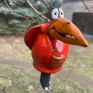 Könnte beinhalten: Handgefertigter Keramik-Vogelanhänger. Der Vogel hat einen leuchtend orangefarbenen Körper, einen langen Schnabel und große, ausdrucksstarke Augen. Er hat eine kleine Tasche am Bauch und einen schwarzen Fuß mit weißen Zehen. Aufgehängt an einer grünen Schnur.