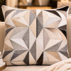 May include: A square throw pillow featuring a geometric patchwork design made of hair-on-hide leather panels in shades of cream, beige, grey, and dark brown. The individual triangular and diamond-shaped pieces are joined with visible stitching.