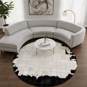May include: A large, light gray curved sectional sofa with a rounded back, paired with a round white marble coffee table featuring gold legs. A circular area rug with a patchwork pattern of white, gray, and black cowhide sits underneath.