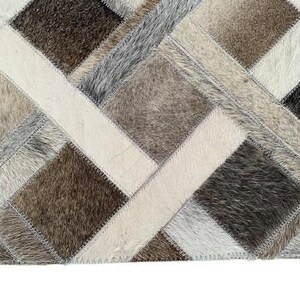 May include: A patchwork rug with a geometric design. The rug features a variety of neutral colors, including cream, gray, brown, and black. The rug is made of a textured material, likely hide or fur, and has a stitched border.