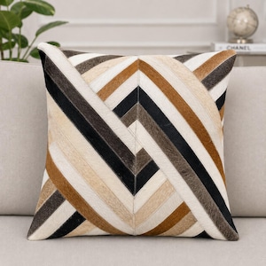 May include: A square throw pillow featuring a geometric chevron pattern composed of stitched strips of hair-on hide. The design displays a color palette of black, dark brown, tan, beige, and white, arranged in alternating diagonal stripes.