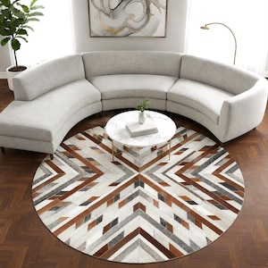Op de afbeelding: Een groot, rond vloerkleed met een geometrisch patroon van bruine, grijze en witte leren stukken. Het kleed ligt onder een ronde salontafel van wit marmer en een lichtgrijze, gebogen hoekbank in een woonkamer.