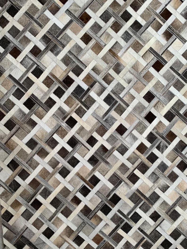 May include: A woven rug with a geometric pattern. The rug features a mix of neutral colors, including shades of brown, gray, and cream. The design is a repeating pattern of interwoven strips, creating a textured surface. The rug is likely made of natural materials.