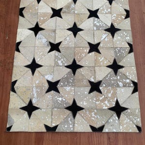 May include: A rectangular area rug constructed from stitched cowhide patches. The design features a repeating geometric pattern of light beige and metallic silver speckled hide segments interspersed with solid black four-pointed star shapes.