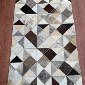 May include: A rectangular area rug constructed from stitched cowhide patches in a geometric pattern. The rug features a variety of triangular and diamond-shaped pieces in shades of white, cream, grey, brown, and black with visible stitching.