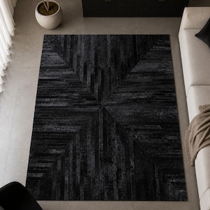 May include: A rectangular area rug featuring a dark charcoal and black color palette with a geometric pattern composed of linear, wood-like slats arranged in a symmetrical, radiating chevron design that creates a textured, layered visual effect.