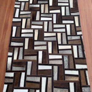 Puede incluir: Una alfombra rectangular construida a partir de un mosaico de piezas de piel rectangulares en varios tonos de marrón, blanco, negro y gris. Las piezas están dispuestas en un patrón geométrico de tejido de cesta con costuras visibles entre cada segmento.