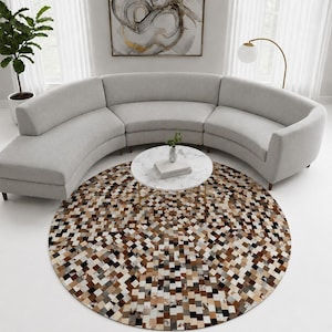 May include: A large, circular area rug featuring a patchwork design of small, square-shaped pieces in various shades of brown, black, white, and gray. The rug is positioned on a white floor in front of a curved, light gray upholstered sofa.