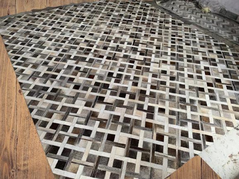 May include: A large, rectangular rug with a woven pattern of alternating light and dark neutral tones. The rug's design features a repeating grid-like structure, creating a textured surface. The color palette includes shades of gray, brown, and cream.