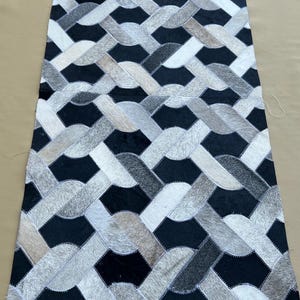 Puede incluir: Alfombra rectangular con un patrón geométrico. La alfombra presenta un diseño tejido con secciones alternas de negro, blanco y varias tonalidades de gris. La alfombra está hecha de un material suave y texturizado, probablemente piel de animal.