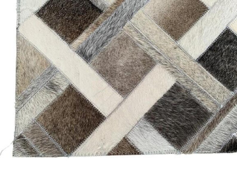 May include: A patchwork rug with a geometric design. The rug features a variety of neutral colors, including cream, gray, and brown. The rug is made of a textured material, possibly cowhide, and has a stitched border.
