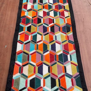 May include: A rectangular area rug featuring a repeating geometric pattern of multicolored hexagons in shades of orange, teal, white, purple, yellow, and black, bordered by a solid black frame made of stitched hair-on-hide leather panels.