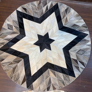 Può includere: Un tappeto circolare composto da singoli pezzi di pelle bovina disposti in un motivo geometrico. Il design presenta una stella nera centrale circondata da strati di tasselli triangolari bianchi, grigi e marroni a raggiera.