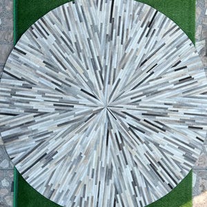 Può includere: Un tappeto rotondo con un design radiale. Il tappeto è realizzato con pezzi rettangolari di pelle nei toni del grigio, bianco e nero, disposti a raggiera dal centro. Il diametro del tappeto è di circa 152 cm.
