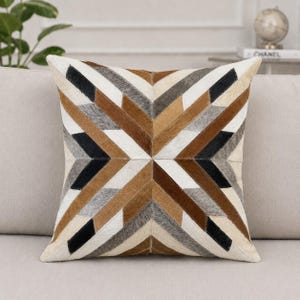 May include: A square throw pillow featuring a geometric pattern made of stitched panels of hair-on hide. The design consists of radiating chevron stripes in alternating colors of cream, tan, dark brown, black, and grey, creating a symmetrical star.