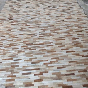 May include: A rectangular rug with a brick-like pattern. The rug features a mix of light beige, tan, and brown rectangular shapes, creating a textured surface. The rug is made of a natural material.