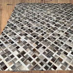 May include: A rectangular rug with a woven pattern of alternating light and dark neutral tones, including shades of gray, brown, and cream. The rug's design features a repeating geometric pattern, creating a textured appearance. The rug is placed on a wooden surface.