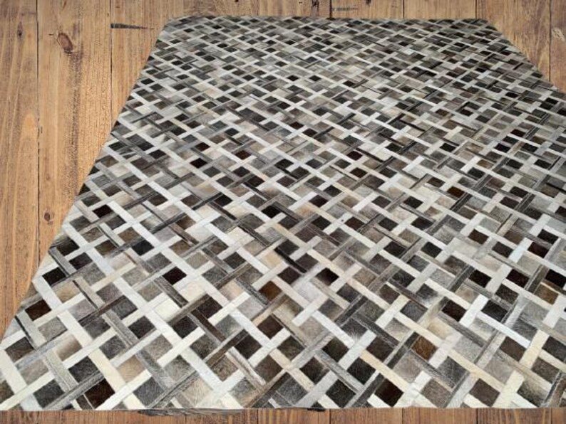 May include: A rectangular rug with a woven pattern of alternating light and dark neutral tones, including shades of gray, brown, and cream. The rug's design features a repeating geometric pattern, creating a textured appearance. The rug is placed on a wooden surface.