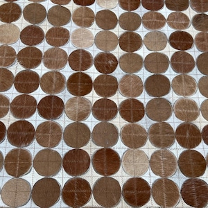 Puede incluir: Una vista de cerca de una alfombra decorativa con un patrón repetitivo de formas circulares marrones. Los círculos están dispuestos en forma de cuadrícula sobre un fondo blanco. La alfombra tiene un aspecto texturizado, con variaciones en los tonos marrones.