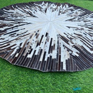 Puede incluir: Alfombra redonda con diseño radial. Presenta un patrón central en forma de estrella en blanco y crema, rodeado de bandas alternas en marrón oscuro y negro. La alfombra está hecha de un material texturizado, probablemente cuero.