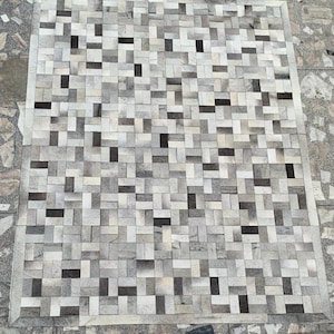 May include: A rectangular area rug constructed from small, rectangular patches of hair-on hide arranged in a staggered, brick-like pattern. The rug features a neutral colour palette consisting of various shades of white, grey, charcoal, and black.