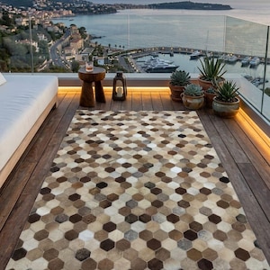 Peut inclure: Un tapis rectangulaire avec un motif géométrique de formes hexagonales marron, beige et crème. Le tapis est posé sur une terrasse en bois, avec une vue sur la mer et une ville en arrière-plan. Le tapis est fait d'une matière douce.