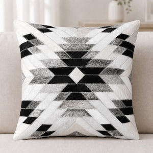 May include: A square throw pillow featuring a geometric pattern constructed from stitched panels of hair-on hide. The design displays a symmetrical arrangement of black, white, and gray sections forming a large central diamond and radiating star.