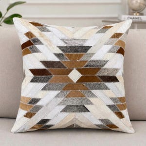 Pode incluir: Uma almofada quadrada com um padrão geométrico de patchwork feito de painéis de couro com pelo. O design consiste em tiras alternadas em tons de branco, creme, bege, castanho e cinzento-carvão, dispostas num motivo de estrela simétrico.