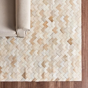 Peut inclure: Un tapis rectangulaire présentant un motif à chevrons tissé composé de carreaux de cuir individuels dans des tons crème, beige et brun clair. Le tapis est placé sous le coin d'un meuble rembourré de couleur beige clair.