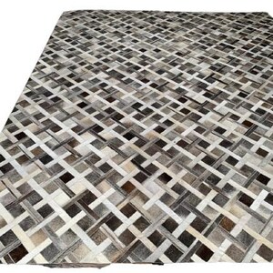 May include: A large, rectangular rug with a woven pattern. The rug features a mix of neutral colors, including white, beige, gray, and brown. The design is a repeating geometric pattern, creating a textured appearance. The rug is made of a soft material.