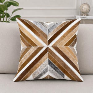 May include: A square throw cushion featuring a geometric chevron pattern made of stitched panels of hair-on hide. The design incorporates alternating diagonal stripes in shades of brown, tan, grey, and white, meeting at a central point.