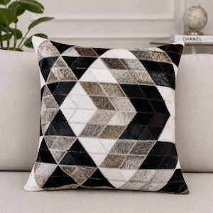 May include: A square throw pillow featuring a geometric patchwork design made of cowhide panels. The pattern consists of repeating triangles and diamond shapes in a color palette of solid black, stark white, and variegated shades of brown and gray.
