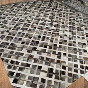 May include: A large, rectangular rug with a woven pattern of alternating light and dark neutral tones. The rug's design features a repeating grid-like structure, creating a textured surface. The color palette includes shades of gray, brown, and cream.