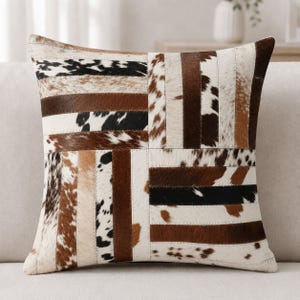 May include: A square throw pillow featuring a patchwork design of cowhide strips. The pattern consists of alternating vertical and horizontal panels in shades of brown, white, and black, showcasing natural animal hide textures and varied fur markings.