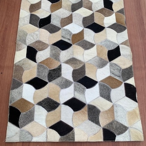 May include: A rectangular area rug constructed from individual patches of cowhide leather. The rug features a repeating geometric pattern of interlocking cubes in a palette of black, white, cream, tan, and various shades of brown and gray.