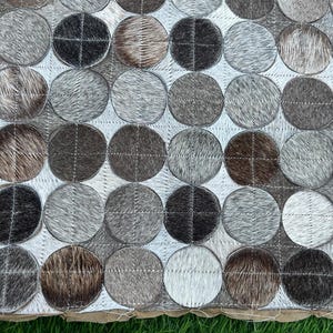 Peut inclure: Un tapis décoratif avec un motif de cercles cousus et texturés dans des tons de gris, de marron et de noir. Les cercles sont disposés en grille sur un fond blanc, créant un design géométrique moderne.
