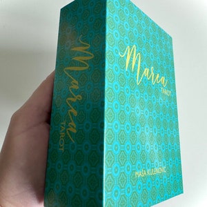 May include: A teal green box with a gold geometric pattern and the text "Marcea Tarot" in gold lettering. The box is designed by Masa Kulenović.