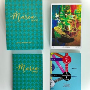 May include: A teal blue box with the text "Marea Tarot" in gold lettering. The box is decorated with a repeating pattern of small teal blue and green squares. The box is sitting on a white surface. A tarot card with a colorful image of a person standing on a platform with a tiger is visible in the background. The card is labeled "0 The Fool".
