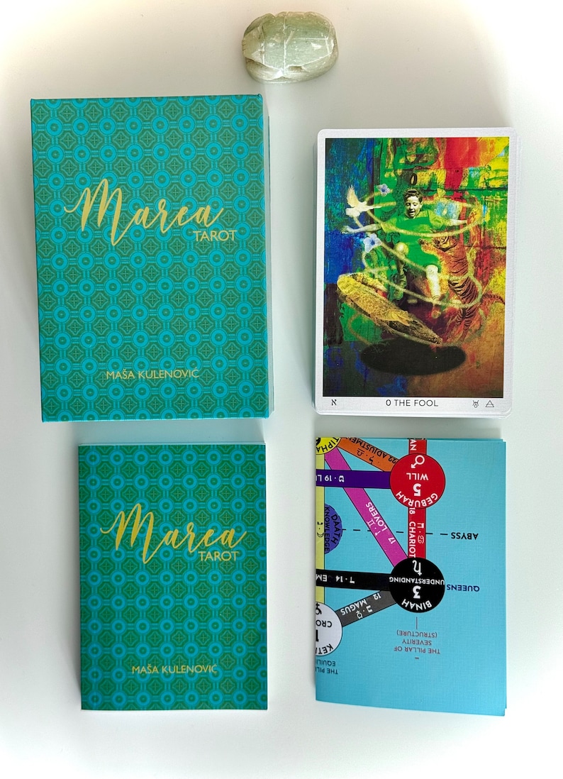 May include: A teal blue box with the text "Marea Tarot" in gold lettering. The box is decorated with a repeating pattern of small teal blue and green squares. The box is sitting on a white surface. A tarot card with a colorful image of a person standing on a platform with a tiger is visible in the background. The card is labeled "0 The Fool".