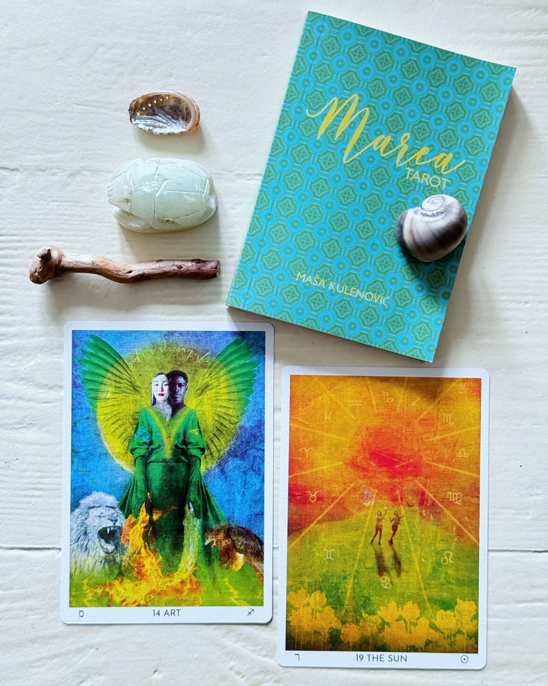 May include: Two tarot cards, one depicting a figure with wings and a lion and wolf, labeled "14 ART", and the other depicting a sunset with two figures, labeled "19 THE SUN". The cards are on a white wooden surface with a book titled "Marra Tarot" by Masa Kulenović, a seashell, and two small decorative stones.