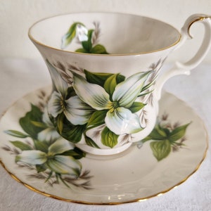 May include: A white teacup and saucer set with a floral design featuring white flowers with green leaves. The set has a gold trim around the edges.
