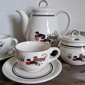 Puede incluir: Un juego de té blanco con un diseño de patos y juncos. El juego incluye una tetera, un azucarero, una jarra de crema y dos tazas y platillos.