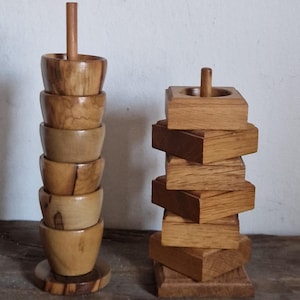 May include: Two wooden egg cups, one stacked with seven cups and the other stacked with six square blocks. Both are made of light brown wood with a natural grain pattern.