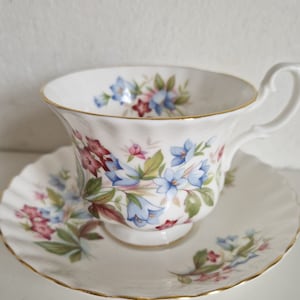 May include: A white teacup and saucer with a floral pattern. The pattern features blue, pink, and green flowers on a white background. The cup and saucer have a gold trim.