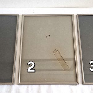 Puede incluir: Tres marcos de fotos plateados con paspartús negros. Los marcos están numerados del 1 al 3. El marco con el número 1 tiene una pegatina verde.