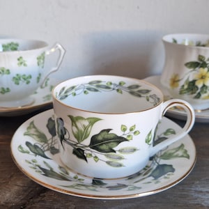 Könnte beinhalten: Drei weiße Teetassen mit Blumenmustern. Die Tassen haben grüne und gelbe Blumenmuster. Die Tassen stehen auf weißen Untertassen mit passenden Blumenmustern.