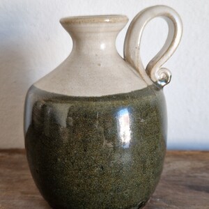 Può includere: Un piccolo boccale in ceramica fatto a mano con corpo arrotondato e un'unica maniglia. Il boccale è smaltato di un colore verde scuro con un bordo e un manico di colore crema più chiaro.