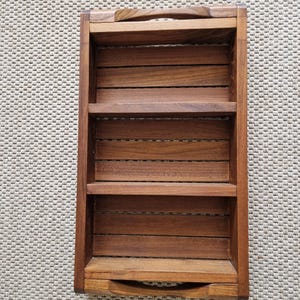 Puede incluir: Una estantería rectangular de madera con tres estantes horizontales y un asa en la parte superior. La estantería es de madera marrón oscuro y tiene un diseño de listones. La estantería está vacía y se encuentra sobre una superficie de color neutro.