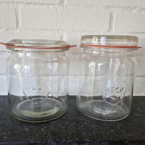 Op de afbeelding: Twee glazen potten met rode rubberen afdichtingen en het woord "WECK" erop gegraveerd.