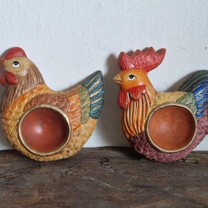 May include: Two ceramic rooster egg holders. One is brown with green, yellow, and blue accents. The other is red, yellow, and blue with a brown base.